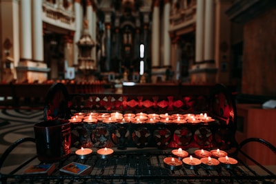 A dimly lit church interior with a foreground focus on small, lit candles placed on a patterned metal stand. The background shows ornate architectural details and large pillars, giving a sense of depth. A mug and matchboxes are visible near the candles.