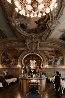 An opulent, intricately decorated dining room features an ornate ceiling with detailed paintings and elaborate molding. A grand chandelier hangs centrally, illuminating the bar area stocked with various bottles and floral arrangements. The room has wooden flooring and classical pillars, with beautifully set tables in the background. A waiter dressed in formal attire is seen walking towards the bar.