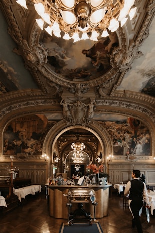 An opulent, intricately decorated dining room features an ornate ceiling with detailed paintings and elaborate molding. A grand chandelier hangs centrally, illuminating the bar area stocked with various bottles and floral arrangements. The room has wooden flooring and classical pillars, with beautifully set tables in the background. A waiter dressed in formal attire is seen walking towards the bar.