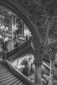 An intricate and grand staircase dominates the scene, adorned with ornate carvings and sculptures. Chandeliers hang elegantly, casting a soft light across the arches and columns of the structure. People in formal attire are seen ascending and descending the stairs, adding a sense of timeless elegance to the environment.