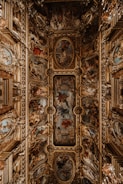 An ornate ceiling features intricate paintings and elaborate gold decorative elements. The artwork is composed of a series of framed frescoes depicting various scenes with figures and rich color contrasts. Surrounding the paintings are three-dimensional, gilded embellishments that add a sense of grandeur and opulence to the space.
