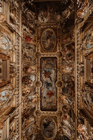 An ornate ceiling features intricate paintings and elaborate gold decorative elements. The artwork is composed of a series of framed frescoes depicting various scenes with figures and rich color contrasts. Surrounding the paintings are three-dimensional, gilded embellishments that add a sense of grandeur and opulence to the space.