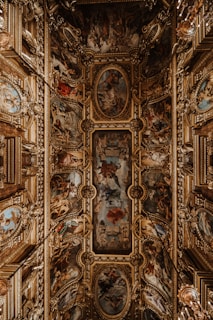 An ornate ceiling features intricate paintings and elaborate gold decorative elements. The artwork is composed of a series of framed frescoes depicting various scenes with figures and rich color contrasts. Surrounding the paintings are three-dimensional, gilded embellishments that add a sense of grandeur and opulence to the space.
