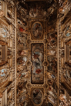 An ornate ceiling features intricate paintings and elaborate gold decorative elements. The artwork is composed of a series of framed frescoes depicting various scenes with figures and rich color contrasts. Surrounding the paintings are three-dimensional, gilded embellishments that add a sense of grandeur and opulence to the space.