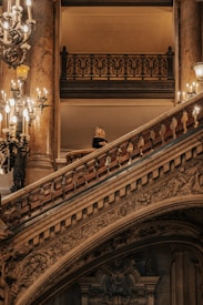 An ornate staircase with intricate carvings and balustrades, illuminated by multiple chandeliers. A person stands on the staircase wearing formal attire, adding a sense of elegance and grandeur to the scene.