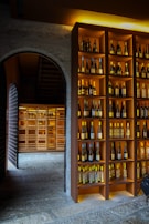 A dimly lit wine cellar featuring shelves filled with various bottles of wine. The shelves are neatly organized with bottles of different colors and sizes, displayed against a warm wooden background. An arched stone doorway leads to another section of the cellar, which is also filled with more bottles, creating a cozy and inviting atmosphere.