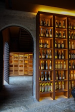 A dimly lit wine cellar featuring shelves filled with various bottles of wine. The shelves are neatly organized with bottles of different colors and sizes, displayed against a warm wooden background. An arched stone doorway leads to another section of the cellar, which is also filled with more bottles, creating a cozy and inviting atmosphere.