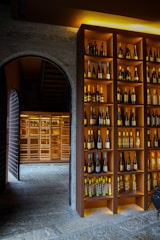 A dimly lit wine cellar featuring shelves filled with various bottles of wine. The shelves are neatly organized with bottles of different colors and sizes, displayed against a warm wooden background. An arched stone doorway leads to another section of the cellar, which is also filled with more bottles, creating a cozy and inviting atmosphere.