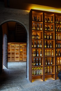 A dimly lit wine cellar featuring shelves filled with various bottles of wine. The shelves are neatly organized with bottles of different colors and sizes, displayed against a warm wooden background. An arched stone doorway leads to another section of the cellar, which is also filled with more bottles, creating a cozy and inviting atmosphere.