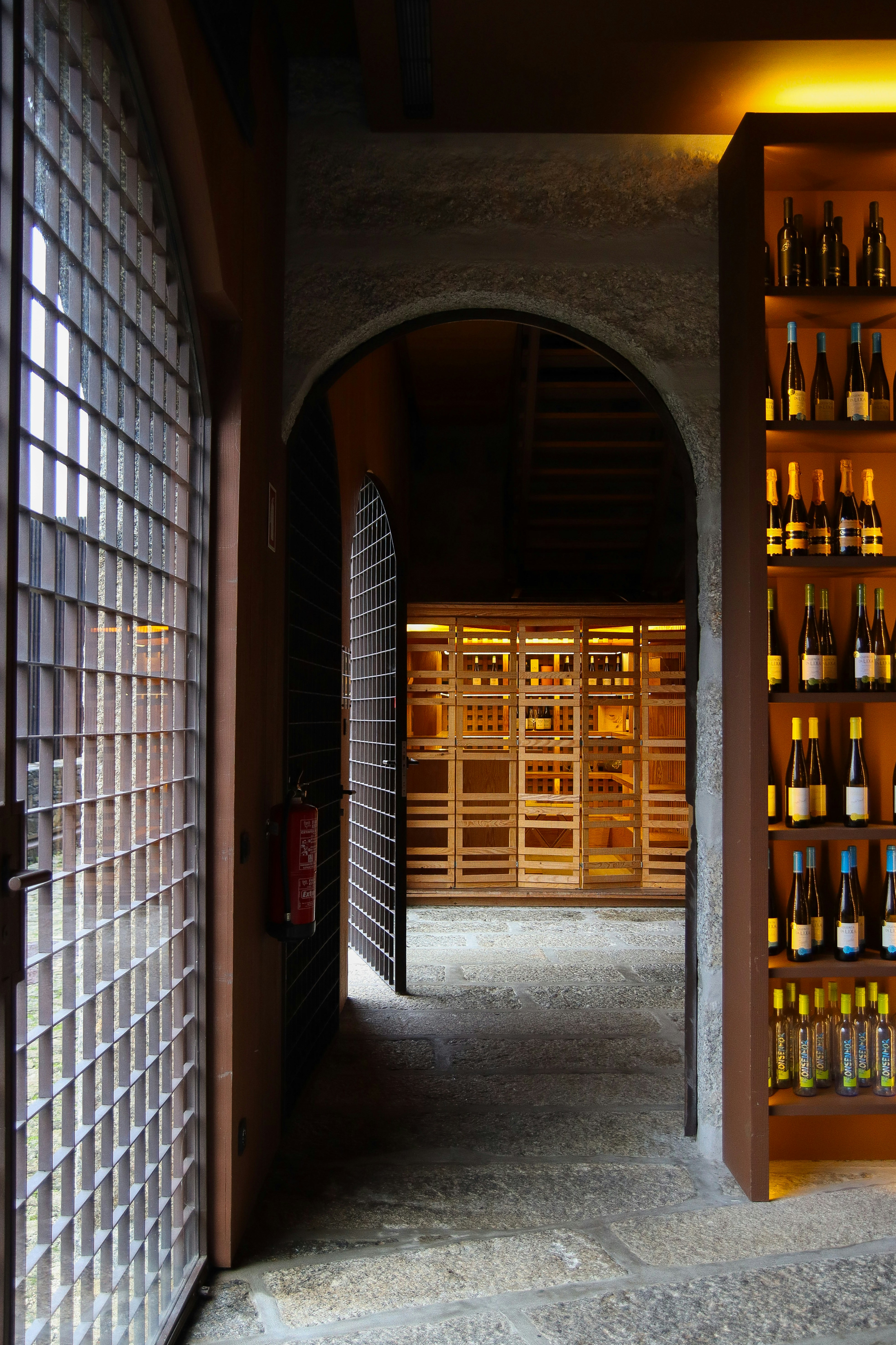 A wine cellar with stone floors, arched entryways, gated doors, and shelves of wine bottles lit with warm orange light.