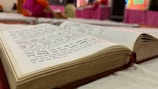 An open book with text in Hindi, lying on a white fabric surface. The background is slightly blurred, suggesting an indoor setting with bright colors, where people are possibly sitting or engaging in an activity.