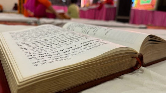 An open book with text in Hindi, lying on a white fabric surface. The background is slightly blurred, suggesting an indoor setting with bright colors, where people are possibly sitting or engaging in an activity.