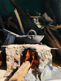 A rustic outdoor cooking setup featuring a metal wok resting on a traditional clay stove. Wooden logs fuel the fire underneath, with flames visible. Surrounding the stove are various wooden sticks and some plastic bags.