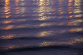 Rippling water with reflections of lights creating a shimmering effect. The lights appear blurred and add a warm, vibrant glow across the gentle waves.