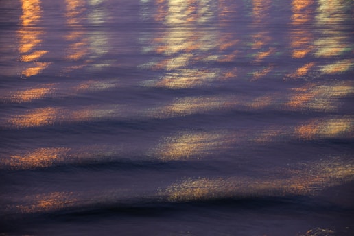 Rippling water with reflections of lights creating a shimmering effect. The lights appear blurred and add a warm, vibrant glow across the gentle waves.