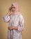 A woman is wearing a pink hijab and a floral-patterned dress with puffed sleeves. Her head is slightly tilted, and her hand touches the side of her scarf. The background is a solid beige color.