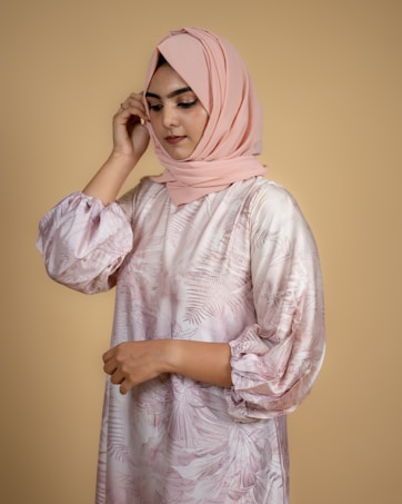 A woman is wearing a pink hijab and a floral-patterned dress with puffed sleeves. Her head is slightly tilted, and her hand touches the side of her scarf. The background is a solid beige color.