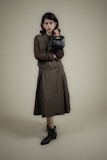A person stands against a plain background wearing a long, dark-colored dress with a matching vest and a textured long-sleeve shirt. The individual holds a small, black handbag in one hand and wears ankle-high black boots. The overall look is formal and stylish.