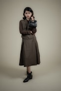 A person stands against a plain background wearing a long, dark-colored dress with a matching vest and a textured long-sleeve shirt. The individual holds a small, black handbag in one hand and wears ankle-high black boots. The overall look is formal and stylish.