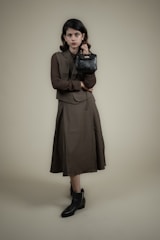 A person stands against a plain background wearing a long, dark-colored dress with a matching vest and a textured long-sleeve shirt. The individual holds a small, black handbag in one hand and wears ankle-high black boots. The overall look is formal and stylish.