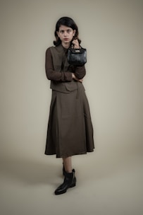 A person stands against a plain background wearing a long, dark-colored dress with a matching vest and a textured long-sleeve shirt. The individual holds a small, black handbag in one hand and wears ankle-high black boots. The overall look is formal and stylish.