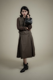 A person stands against a plain background wearing a long, dark-colored dress with a matching vest and a textured long-sleeve shirt. The individual holds a small, black handbag in one hand and wears ankle-high black boots. The overall look is formal and stylish.