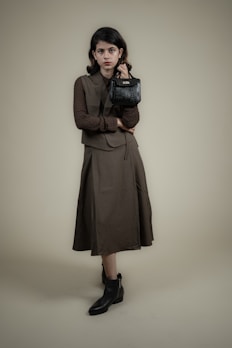 A person stands against a plain background wearing a long, dark-colored dress with a matching vest and a textured long-sleeve shirt. The individual holds a small, black handbag in one hand and wears ankle-high black boots. The overall look is formal and stylish.