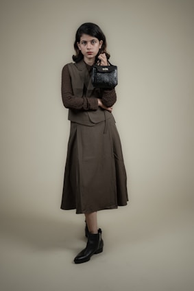 A person stands against a plain background wearing a long, dark-colored dress with a matching vest and a textured long-sleeve shirt. The individual holds a small, black handbag in one hand and wears ankle-high black boots. The overall look is formal and stylish.