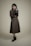 A person stands against a plain background wearing a long, dark-colored dress with a matching vest and a textured long-sleeve shirt. The individual holds a small, black handbag in one hand and wears ankle-high black boots. The overall look is formal and stylish.