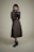 A person stands against a plain background wearing a long, dark-colored dress with a matching vest and a textured long-sleeve shirt. The individual holds a small, black handbag in one hand and wears ankle-high black boots. The overall look is formal and stylish.