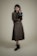 A person stands against a plain background wearing a long, dark-colored dress with a matching vest and a textured long-sleeve shirt. The individual holds a small, black handbag in one hand and wears ankle-high black boots. The overall look is formal and stylish.