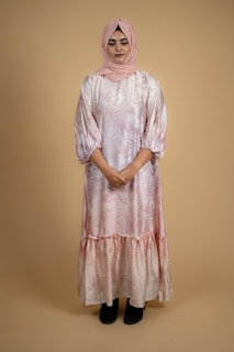 Elegant woman wearing a flowing modest dress in soft pastel tones, standing against a minimal white background.