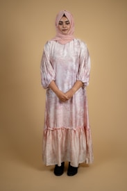 A person is standing against a plain beige background, wearing a long, flowing dress with a subtle pattern in shades of pink. The dress has long sleeves and is paired with a matching pink headscarf. The person is standing still with arms gently folded, and wearing dark footwear.