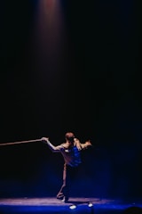 A performer stands under a spotlight on a dark stage, holding a rope in each hand. The individual is dressed in a formal outfit and strikes a dynamic pose, creating an engaging visual contrast against the dimly lit background.