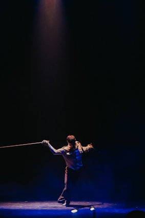 A performer stands under a spotlight on a dark stage, holding a rope in each hand. The individual is dressed in a formal outfit and strikes a dynamic pose, creating an engaging visual contrast against the dimly lit background.
