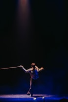 A performer stands under a spotlight on a dark stage, holding a rope in each hand. The individual is dressed in a formal outfit and strikes a dynamic pose, creating an engaging visual contrast against the dimly lit background.