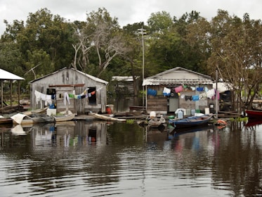 Two rustic wooden houses are situated on the edge of a body of water, with laundry hanging across lines strung between them. Small wooden boats are moored nearby, floating gently on the calm, reflective water. Lush green trees surround the area, creating a serene backdrop.