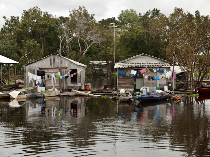 Two rustic wooden houses are situated on the edge of a body of water, with laundry hanging across lines strung between them. Small wooden boats are moored nearby, floating gently on the calm, reflective water. Lush green trees surround the area, creating a serene backdrop.