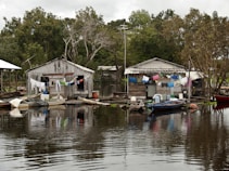Two rustic wooden houses are situated on the edge of a body of water, with laundry hanging across lines strung between them. Small wooden boats are moored nearby, floating gently on the calm, reflective water. Lush green trees surround the area, creating a serene backdrop.