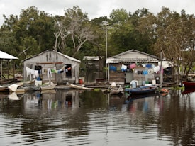 Two rustic wooden houses are situated on the edge of a body of water, with laundry hanging across lines strung between them. Small wooden boats are moored nearby, floating gently on the calm, reflective water. Lush green trees surround the area, creating a serene backdrop.