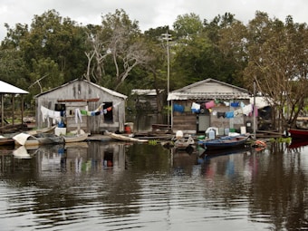 Two rustic wooden houses are situated on the edge of a body of water, with laundry hanging across lines strung between them. Small wooden boats are moored nearby, floating gently on the calm, reflective water. Lush green trees surround the area, creating a serene backdrop.