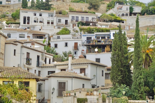 A hillside adorned with white Mediterranean-style houses featuring terracotta roof tiles. Several homes have small balconies with plants and satellite dishes. Tall cypress trees and a palm tree add greenery, while the background consists of more houses and a rustic stone wall.