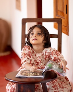 A young child in a floral-patterned dress is sitting on a wooden chair, holding and looking at an open book placed on a round wooden table. The child appears to be gazing upwards with a thoughtful expression. The background features a red floor, part of a wall with a window, and soft natural lighting coming from the side.