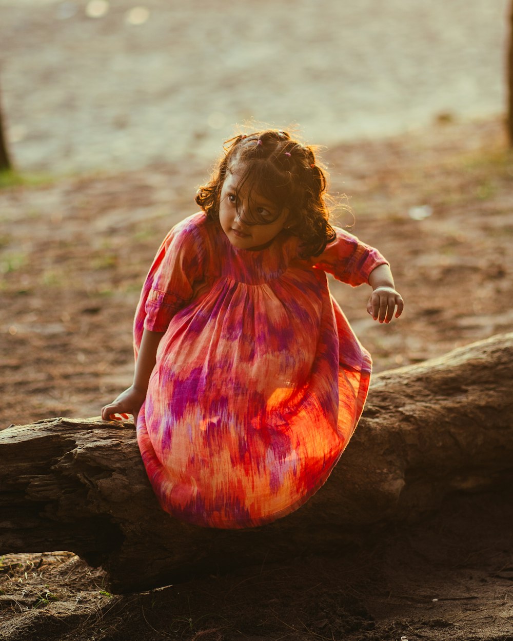 a little girl in a colorful dress sitting on a log