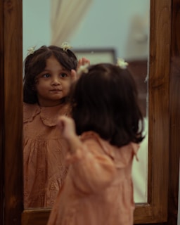 A young girl in a peach-colored dress is looking at her reflection in a wooden-framed mirror. Small flowers adorn her hair, and she appears to be intently observing her own expression. The background is softly blurred, with neutral tones.