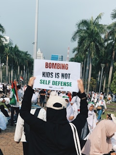 A group of people gathered outdoors, with one person holding up a sign that reads 'Bombing kids is not self defense'. The setting is a palm tree-lined area that resembles a park or public space. Many participants are wearing white, and several are wearing headscarves. The atmosphere appears to be of a protest or rally.