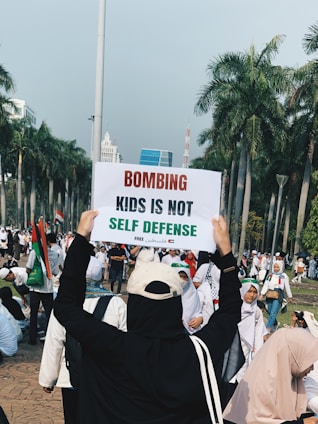 A group of people gathered outdoors, with one person holding up a sign that reads 'Bombing kids is not self defense'. The setting is a palm tree-lined area that resembles a park or public space. Many participants are wearing white, and several are wearing headscarves. The atmosphere appears to be of a protest or rally.