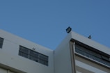 A modern building features large rectangular windows and flat roof infrastructure with mounted lights against a clear blue sky.