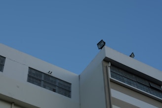 A modern building features large rectangular windows and flat roof infrastructure with mounted lights against a clear blue sky.