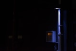 A dimly lit scene featuring a vertical pipe and an electrical panel with glowing indicator lights. The surrounding environment is mostly dark, highlighting the contrast with the illuminated objects.